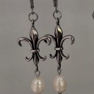 Puede incluir: Pendientes plateados con diseño de flor de lis y una perla colgante. Los pendientes presentan un diseño floral estilizado y detallado y están suspendidos de cierres de palanca. Las perlas son de color blanco crema.