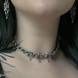 May include: A silver chain choker necklace with a delicate, intricate design. The necklace has small, round pendants hanging from it.