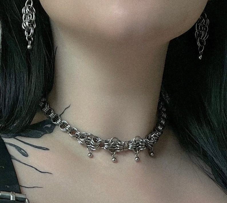 May include: A silver chain choker necklace with a delicate, intricate design. The necklace has small, round pendants hanging from it.