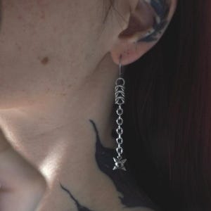 Puede incluir: Un pendiente colgante de tono plateado con un diseño de cadena vertical. La parte superior consta de eslabones circulares entrelazados, mientras que el extremo inferior termina en un pequeño dije en forma de estrella de cuatro puntas con un detalle esférico central.