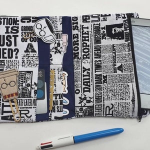 Pode incluir: Capa para leitor de livros eletrónicos com um design de jornal, com um bolso com zíper. A capa tem vários bolsos e alfinetes decorativos. Uma caneta azul e branca está em primeiro plano.