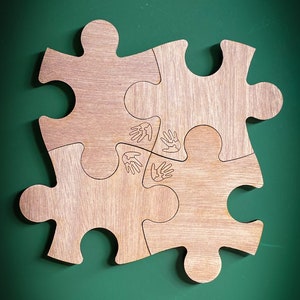 Pode incluir: Três peças de quebra-cabeça de madeira encaixam-se para formar uma peça de quebra-cabeça maior. O centro do quebra-cabeça tem uma imagem de quatro mãos entrelaçadas.