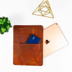 Könnte beinhalten: Eine braune Lederhülle für ein Tablet mit einer Tasche für ein Telefon. Die Hülle trägt den Namen "Oliver" geprägt auf der Vorderseite. Ein blaues Telefon befindet sich in der Tasche.