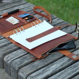 May include: A brown leather organizer with a notepad, pen, and pockets for cards and cash. The organizer has a zipper closure and a wrist strap. A smartphone is visible inside the zippered pocket. Sunglasses rest on the wooden surface.