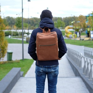 May include: A person wearing a blue jacket and jeans is carrying a brown leather backpack with a zippered pocket.