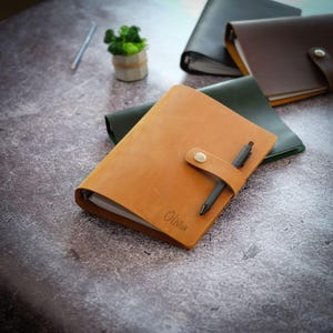 Puede incluir: Una carpeta de cuero color canela con una correa de cierre de botón a presión, un bolígrafo negro insertado en la correa y el nombre Olivia grabado en la portada. La carpeta contiene páginas de papel blanco y es parte de una colección similar.