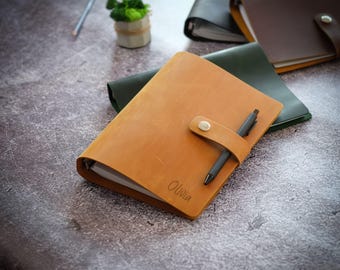 Puede incluir: Una carpeta de cuero color canela con una correa de cierre de botón a presión, un bolígrafo negro insertado en la correa y el nombre Olivia grabado en la portada. La carpeta contiene páginas de papel blanco y es parte de una colección similar.