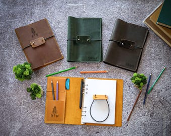 Puede incluir: Tres cuadernos de cuero en marrón, verde y marrón oscuro, cada uno con un cierre elástico negro y una lengüeta de cuero. Un cuaderno de cuero color canela abierto muestra una anilla de seis aros, papel blanco, un bolígrafo y bolsillos.
