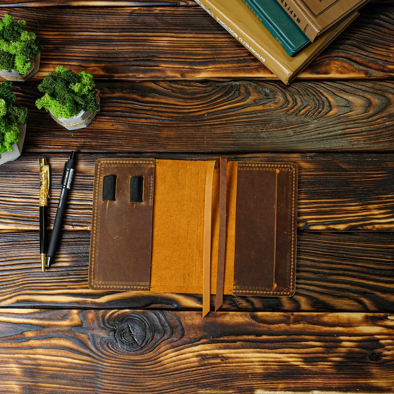 May include: Open brown leather journal with a tan interior, two pen holders, and two brown ribbon bookmarks. The journal is on a dark wooden surface, with two pens and small potted plants with green moss.