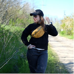 May include: A black baseball cap, a black long-sleeved sweatshirt, and a mustard yellow fanny pack with the name Emily embossed on the front. The fanny pack features a silver zipper closure and is worn across the chest over the black sweatshirt.