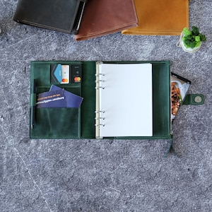 Puede incluir: Una agenda de anillas de cuero verde oscuro con un mecanismo met&aacute;lico de seis anillas, varias ranuras para tarjetas, un portal&aacute;pices con un bol&iacute;grafo negro y un bolsillo lateral con cremallera. La agenda est&aacute; abierta y muestra p&aacute;ginas blancas.