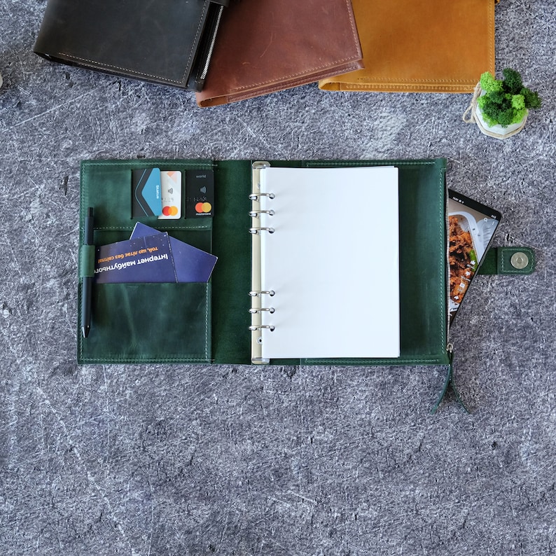 Puede incluir: Una agenda de anillas de cuero verde oscuro con un mecanismo met&aacute;lico de seis anillas, varias ranuras para tarjetas, un portal&aacute;pices con un bol&iacute;grafo negro y un bolsillo lateral con cremallera. La agenda est&aacute; abierta y muestra p&aacute;ginas blancas.
