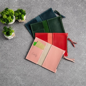 May include: Four leather passport holders in blue, green, red, and pink. Each holder features a side zipper, internal card slots, and visible stitching. The pink holder is personalized with the name Emily engraved on the front pocket.
