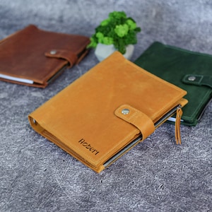Puede incluir: Una funda de cuero color canela para cuaderno con cierre de bot&oacute;n a presi&oacute;n, cremallera lateral y el nombre Robert grabado en la parte frontal. Se muestra junto a un cuaderno marr&oacute;n y uno verde oscuro, ambos con un cierre de bot&oacute;n a presi&oacute;n.