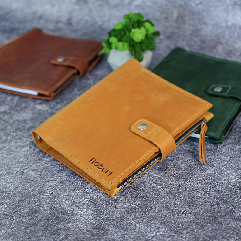 Puede incluir: Una funda de cuero color canela para cuaderno con cierre de bot&oacute;n a presi&oacute;n, cremallera lateral y el nombre Robert grabado en la parte frontal. Se muestra junto a un cuaderno marr&oacute;n y uno verde oscuro, ambos con un cierre de bot&oacute;n a presi&oacute;n.