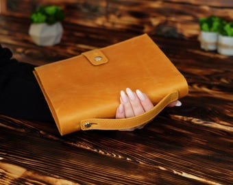 May include: A rectangular tan leather portfolio case featuring a matching leather handle attached with metal rivets and a single snap button closure on the front flap. The exterior surface has a smooth, matte finish with visible natural grain texture.