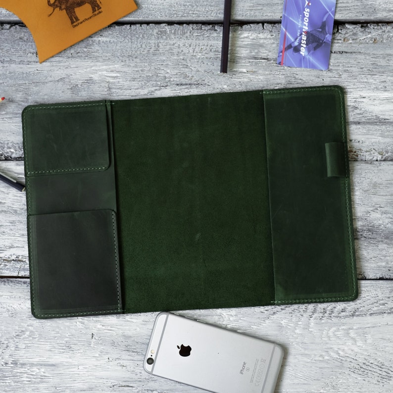 Puede incluir: Carpeta de cuero verde oscuro abierta, con m&uacute;ltiples bolsillos y un portal&aacute;pices. La carpeta est&aacute; abierta sobre una superficie de madera blanca, con un iPhone plateado y una peque&ntilde;a bolsa de cuero con un logotipo de elefante.