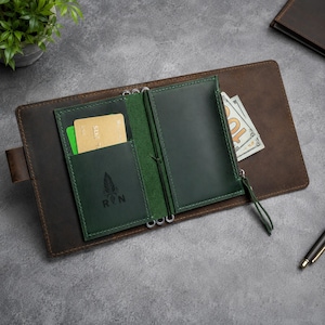 Pode incluir: Uma capa de caderno de couro castanho escuro contendo um inserto de carteira de couro verde escuro. O inserto verde apresenta um compartimento para cartões com um cartão dourado, um bolso com um logótipo de pena e as letras RN.