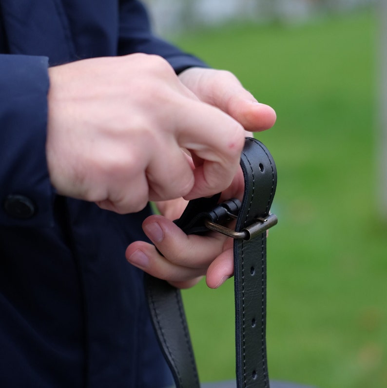 May include: A person adjusts a black leather belt with a silver buckle. The belt has multiple rows of stitching and is made of a durable material.