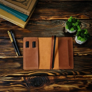 May include: Open brown leather journal with a pen holder and a bookmark. Two pens with gold and black accents are on the left. The journal is on a dark wooden surface. Two small potted plants with green moss are in the background.