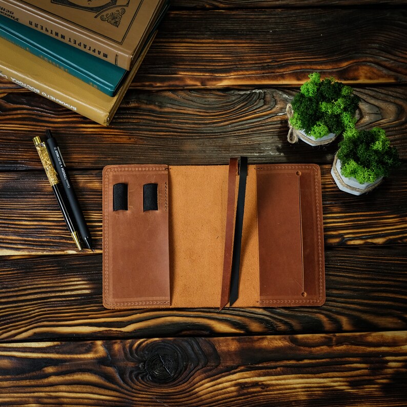 May include: Open brown leather journal with a pen holder and a bookmark. Two pens with gold and black accents are on the left. The journal is on a dark wooden surface. Two small potted plants with green moss are in the background.