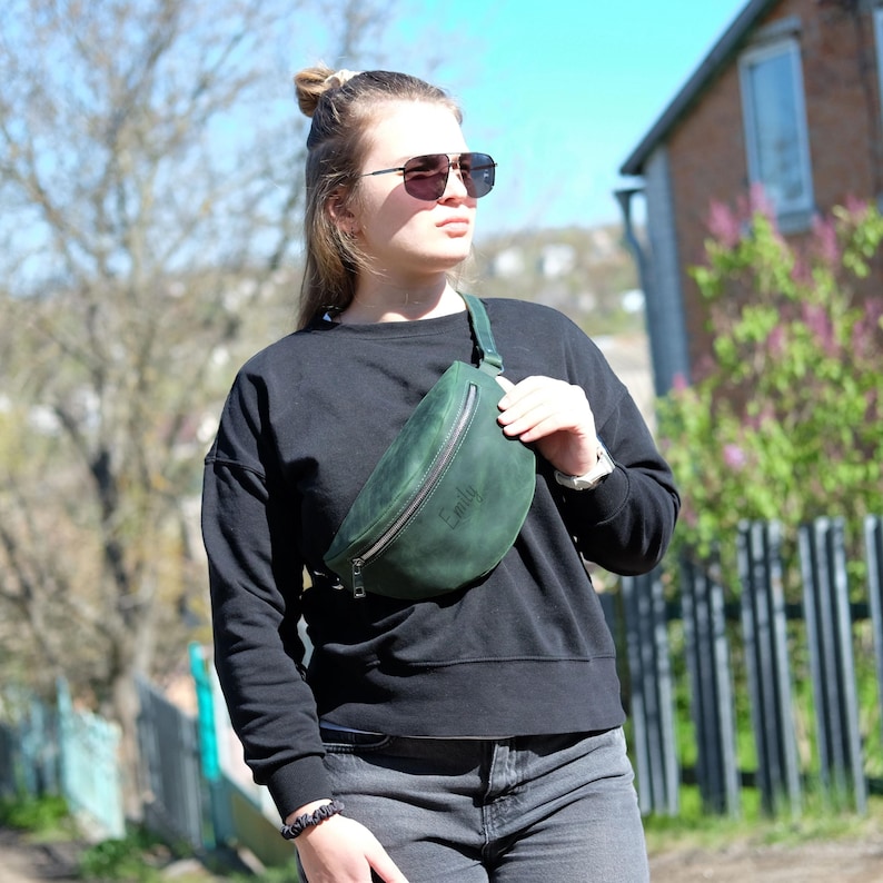 May include: A dark green leather fanny pack featuring a silver zipper closure and the name Emily embossed on the front. The bag is worn across the chest with an adjustable strap, showcasing a smooth matte texture and a curved, crescent-like shape.
