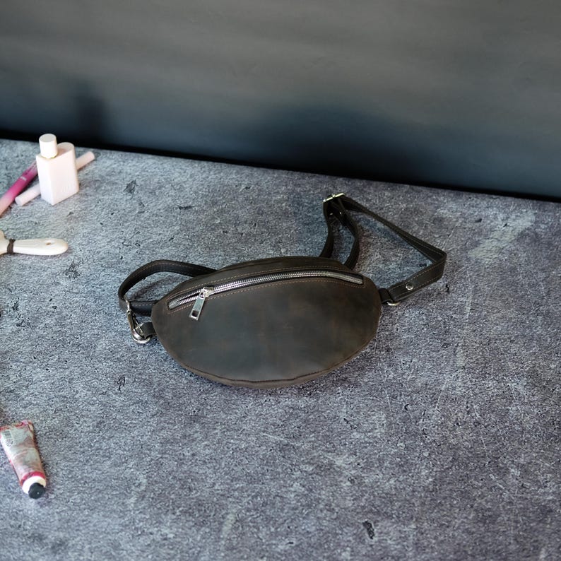May include: A dark brown leather fanny pack featuring a smooth, matte finish and a silver-toned metal zipper closure across the top. The bag includes an adjustable matching leather strap with silver-toned hardware, designed for waist or crossbody wear.