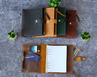 May include: A collection of leather planners in dark brown, tan, green, and reddish-brown, each featuring a snap closure and zipper. One open planner displays a six-ring metal binder mechanism, white paper, card slots, a pen loop, and a black pen.