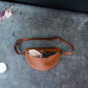 May include: A tan leather fanny pack with an adjustable strap and a zippered main compartment. The bag is open, revealing a white hairbrush and other small items tucked inside. The design features a curved, half-moon shape with visible stitching.