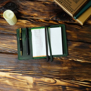 May include: A green leather notebook cover with a grid-lined notepad, two pens, and two dark green ribbon bookmarks. A white candle and books are in the background. The items are on a dark brown wooden surface.