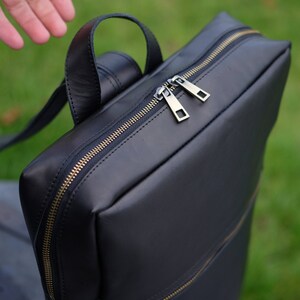 May include: A black leather backpack with a zipper closure and gold accents. The backpack has a top handle and adjustable straps.