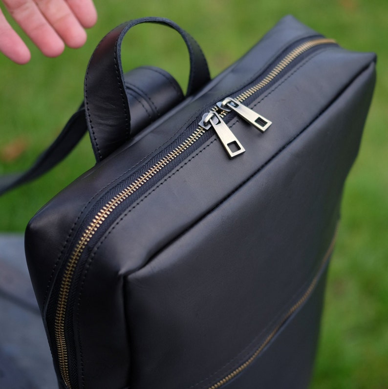 May include: A black leather backpack with a zipper closure and gold accents. The backpack has a top handle and adjustable straps.