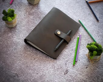 May include: A dark brown leather journal with a snap-button closure strap and a black pen inserted through the loop. The journal is surrounded by several colored pencils and small potted green moss plants on a textured gray surface.