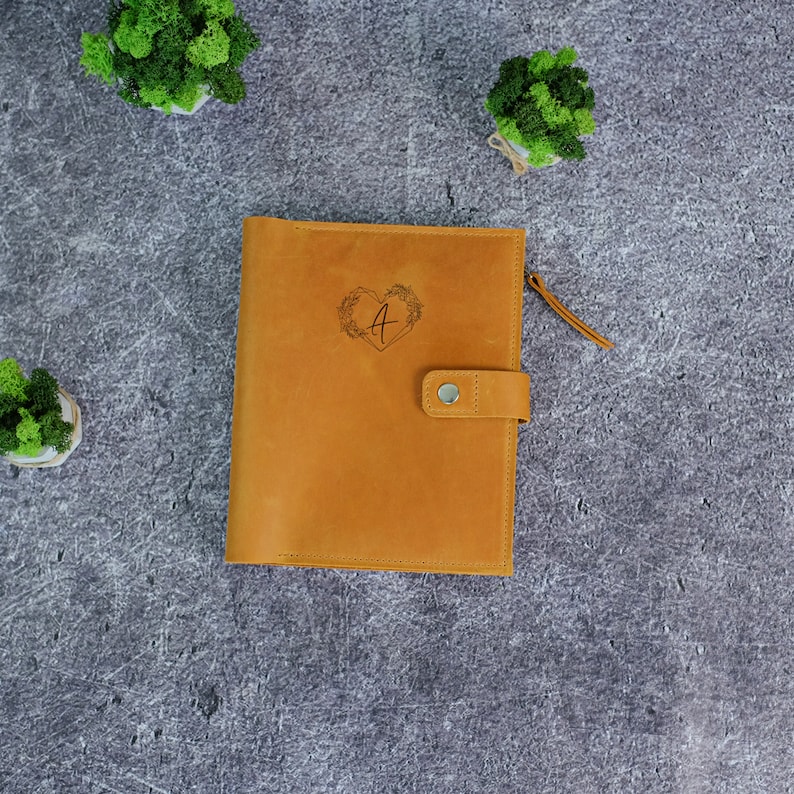 Puede incluir: Un cuaderno de cuero color canela con cierre de broche y una leng&uuml;eta de cuero. La portada presenta un dise&ntilde;o grabado con l&aacute;ser de la letra A centrada dentro de un coraz&oacute;n formado por enredaderas florales. El cuaderno tiene costuras visibles.