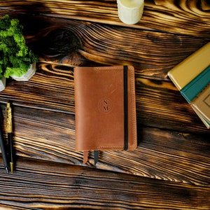 May include: A brown leather journal with a black elastic band closure and a ribbon bookmark. The cover features the initials "N" and "M" inside a circle. The journal is on a wooden surface with other stationery items.