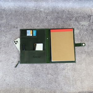 May include: A dark green leather portfolio organizer featuring a snap closure, a notepad with a red top edge, multiple interior card slots, a pen holder, and a pocket containing a white smartphone. The name Oscar is embossed on the front pocket.