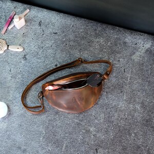 May include: A brown leather fanny pack with an adjustable strap and a silver zipper closure. The front of the bag is embossed with the name Olivia. The interior contains a small patterned item and a pair of dark sunglasses resting inside.