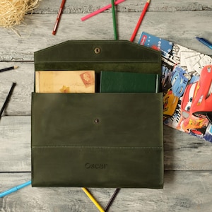 May include: A dark olive green leather envelope-style document holder with a snap button closure and the word Oscar embossed on the front. The interior contains a beige book with a floral illustration and a solid dark green notebook.