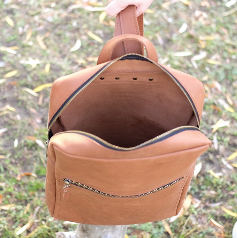May include: A brown leather backpack with a zippered closure and a top handle. The backpack is open and the inside is visible.