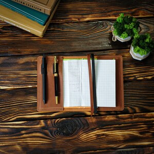 May include: A brown leather notebook with a grid-lined paper interior, open to a partially filled page. Two pens, one black and one gold, are nestled in the cover. A black and a brown ribbon mark the pages. The notebook rests on a dark wooden surface.