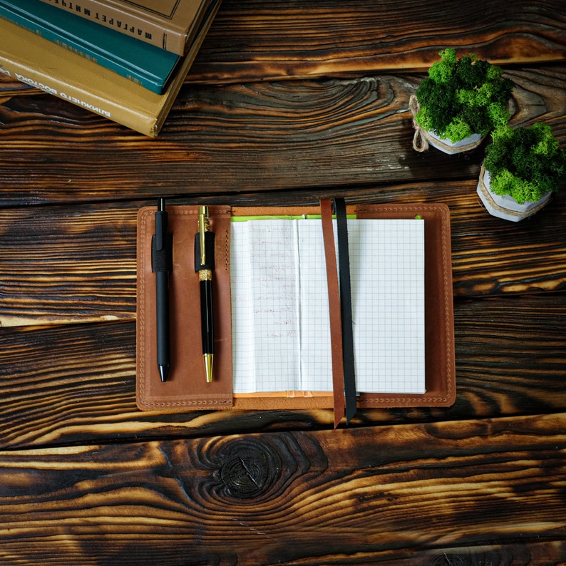 May include: A brown leather notebook with a grid-lined paper interior, open to a partially filled page. Two pens, one black and one gold, are nestled in the cover. A black and a brown ribbon mark the pages. The notebook rests on a dark wooden surface.