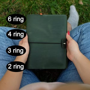 May include: A dark green leather binder featuring a black elastic closure with a metal stud. The image includes text labels indicating options for 6 ring, 4 ring, 3 ring, and 2 ring mechanisms for the planner cover.