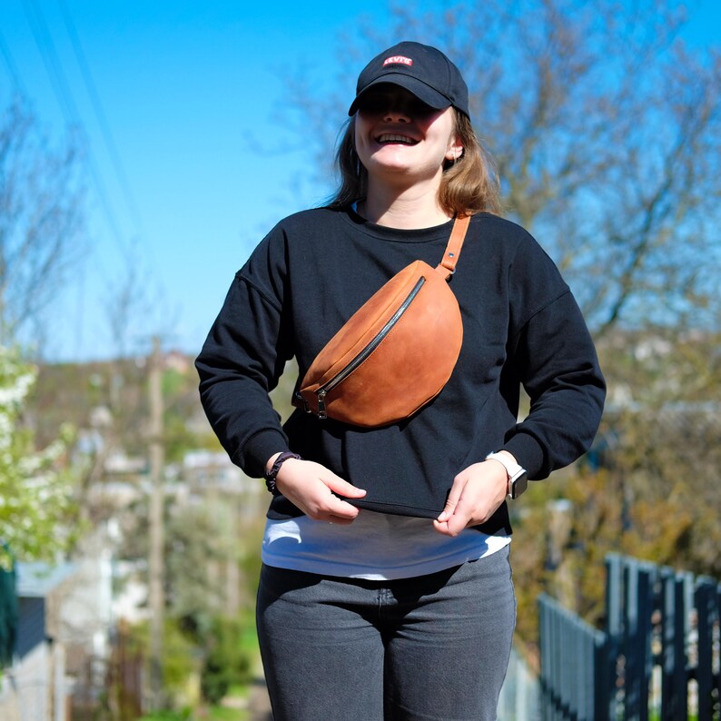 May include: A tan leather fanny pack worn across the chest, featuring a black zipper closure and an adjustable strap. The bag is paired with a black long-sleeve sweatshirt, a white undershirt, dark gray jeans, and a black baseball cap with a red logo.