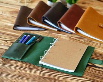 Puede incluir: Una carpeta de cuero verde con un mecanismo de cuatro anillas que sujeta un cuaderno color canela. El interior cuenta con un bolsillo con el nombre Mark grabado, que contiene tres bolígrafos. Otras carpetas de cuero marrón, negro y canela están al fondo.