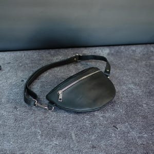 May include: A black fanny pack featuring a smooth, matte finish, a curved silhouette, and a silver-toned metal zipper closure across the top. The bag includes an adjustable black strap with a silver-toned metal clasp and hardware attachments.
