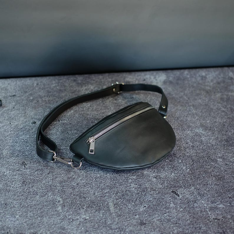 May include: A black fanny pack featuring a smooth, matte finish, a curved silhouette, and a silver-toned metal zipper closure across the top. The bag includes an adjustable black strap with a silver-toned metal clasp and hardware attachments.