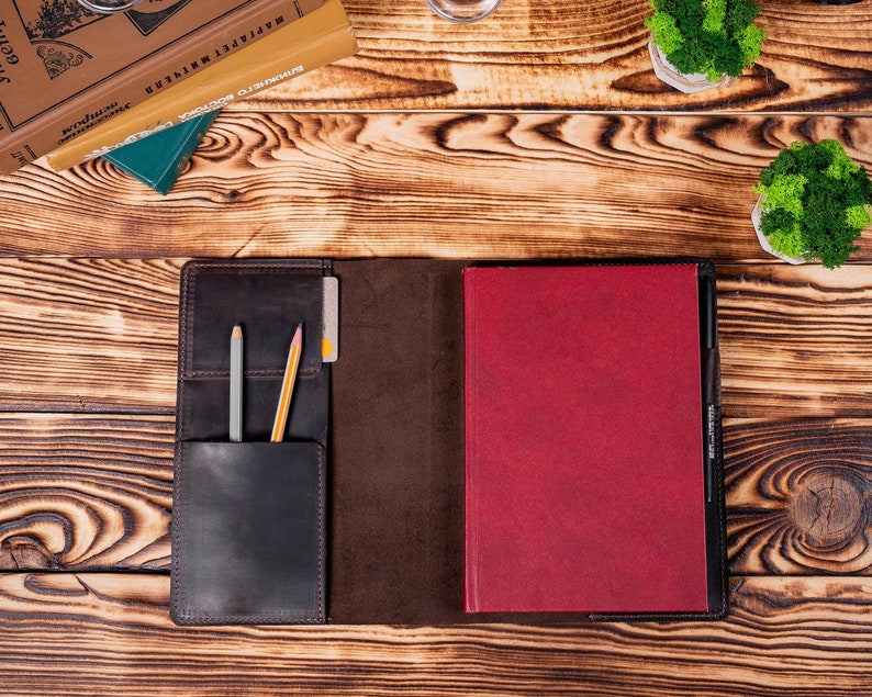 Puede incluir: Funda de cuaderno de cuero abierta con exterior marr&oacute;n oscuro e interior burdeos. Un bolsillo contiene un l&aacute;piz gris, un l&aacute;piz amarillo y una tarjeta de cr&eacute;dito. El cuaderno est&aacute; sobre una superficie de madera con libros y peque&ntilde;as plantas en macetas.