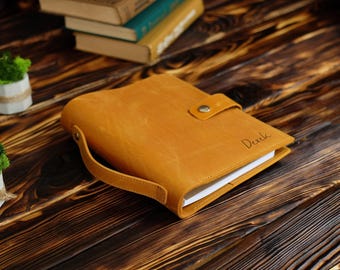 May include: A tan leather notebook with a side handle and a snap button closure. The cover features the name Derek engraved in a cursive script. The notebook contains white paper pages and is positioned horizontally on a dark wooden surface.