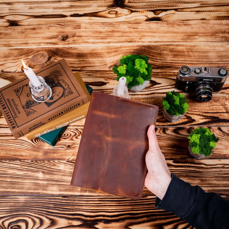 Puede incluir: Un diario de cuero marr&oacute;n se sostiene contra una superficie de madera. El diario est&aacute; rodeado de libros, una vela encendida en un portavelas de vidrio, peque&ntilde;as plantas en macetas y una c&aacute;mara vintage. El fondo de vetas de madera a&ntilde;ade un toque r&uacute;stico.