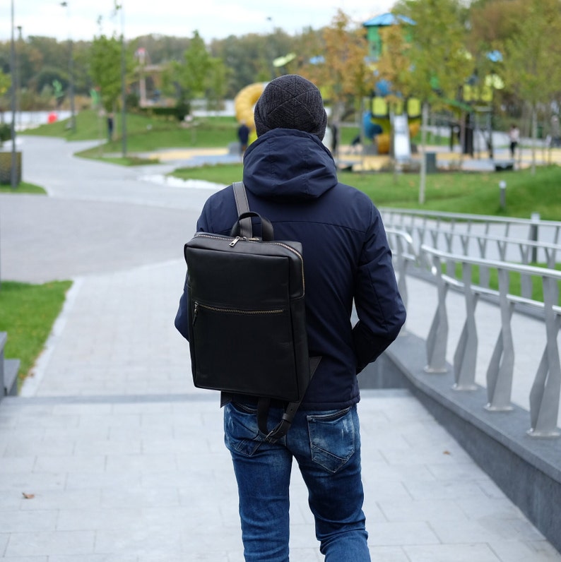 May include: A person wearing a dark blue jacket and jeans is walking away from the camera. They are carrying a black leather backpack with a zippered pocket on the front.